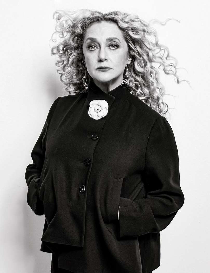 Black and white portrait of an adult female with curly blonde hair wearing a black jacket with a white flower pin against a white background.