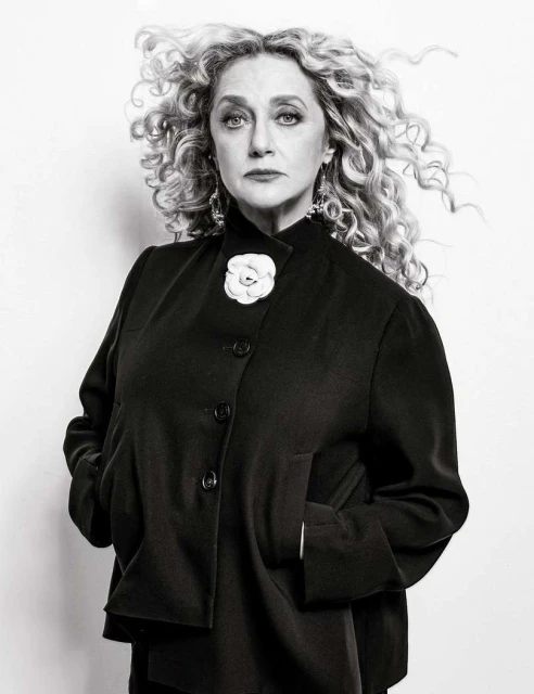 Black and white portrait of an adult female with curly blonde hair wearing a black jacket with a white flower pin against a white background.