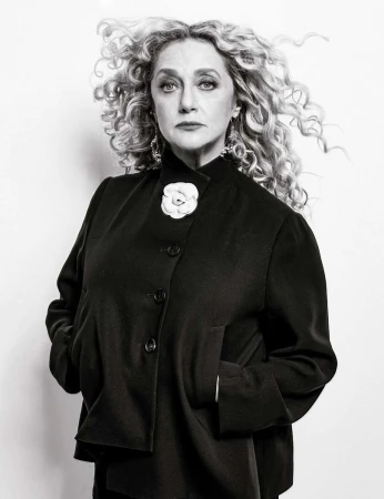Black and white portrait of an adult female with curly blonde hair wearing a black jacket with a white flower pin against a white background.