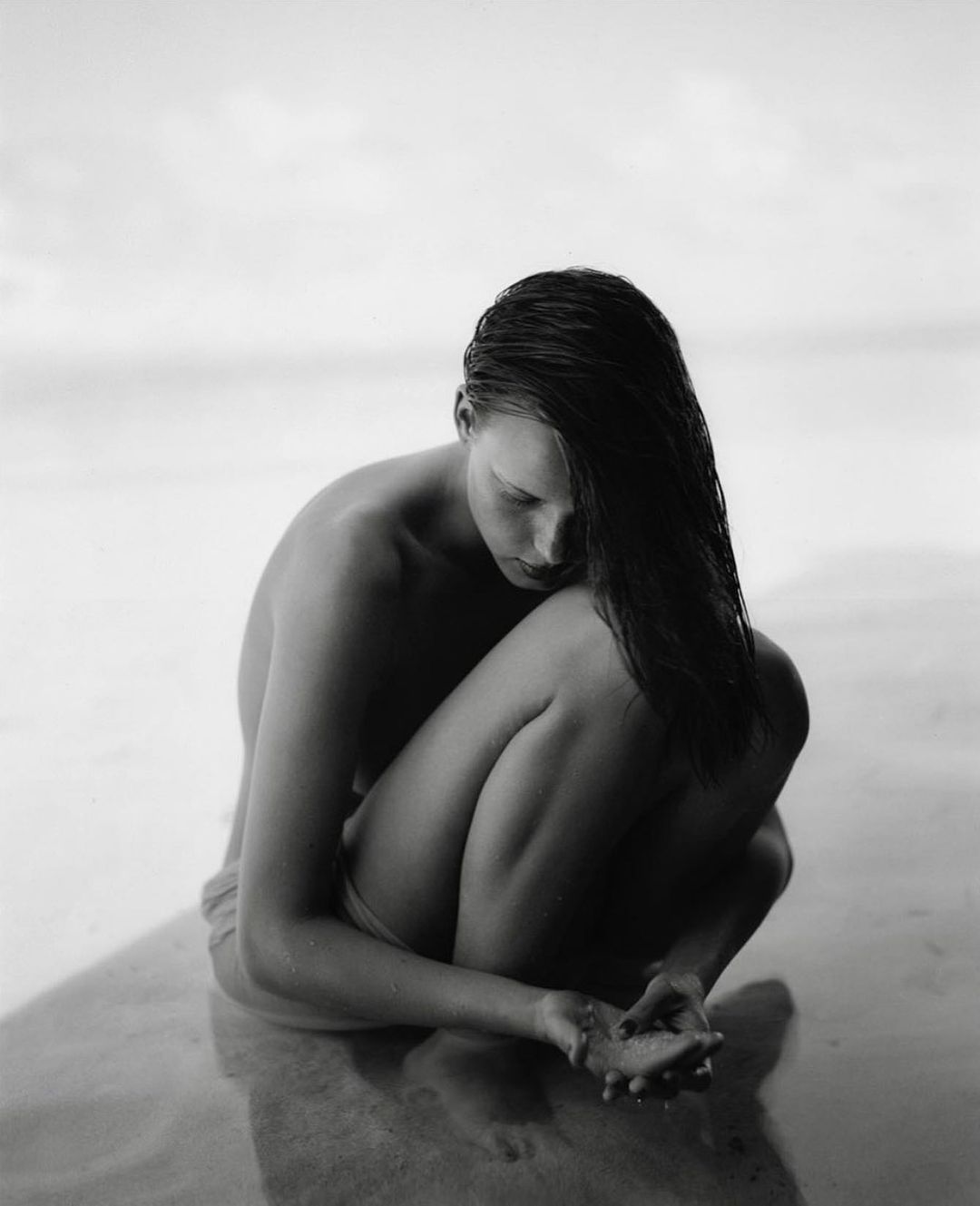 Black and white portrait of a young adult woman sitting at the beach with her legs folded, head down, and wet hair.