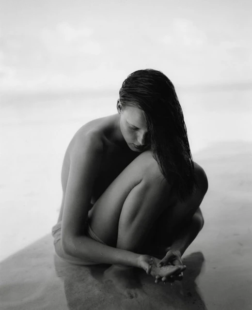 Black and white portrait of a young adult woman sitting at the beach with her legs folded, head down, and wet hair.