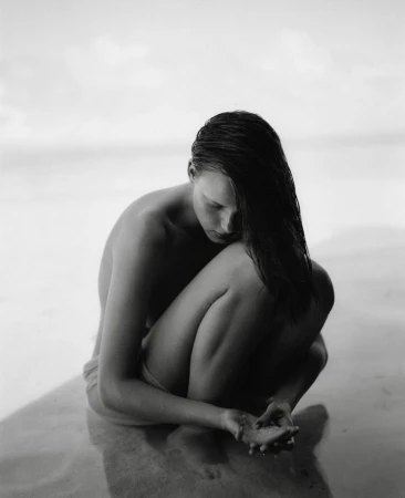 Monochrome portrait of a woman crouched on a sandy beach looking down at her hands holding sand