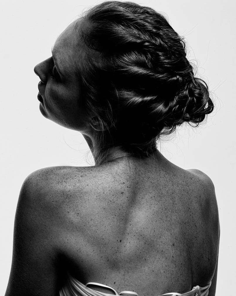 Monochrome portrait of a woman with an intricate braided hairstyle, pale skin, and visible freckles posing against a bright white backdrop.