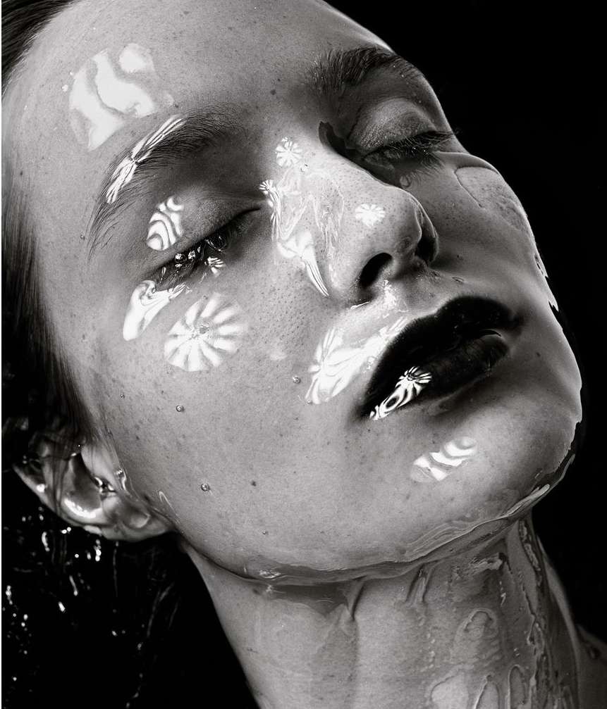 Monochrome close-up of a woman's face with wet skin and black lipstick, featuring patterned light reflections and a dark background.