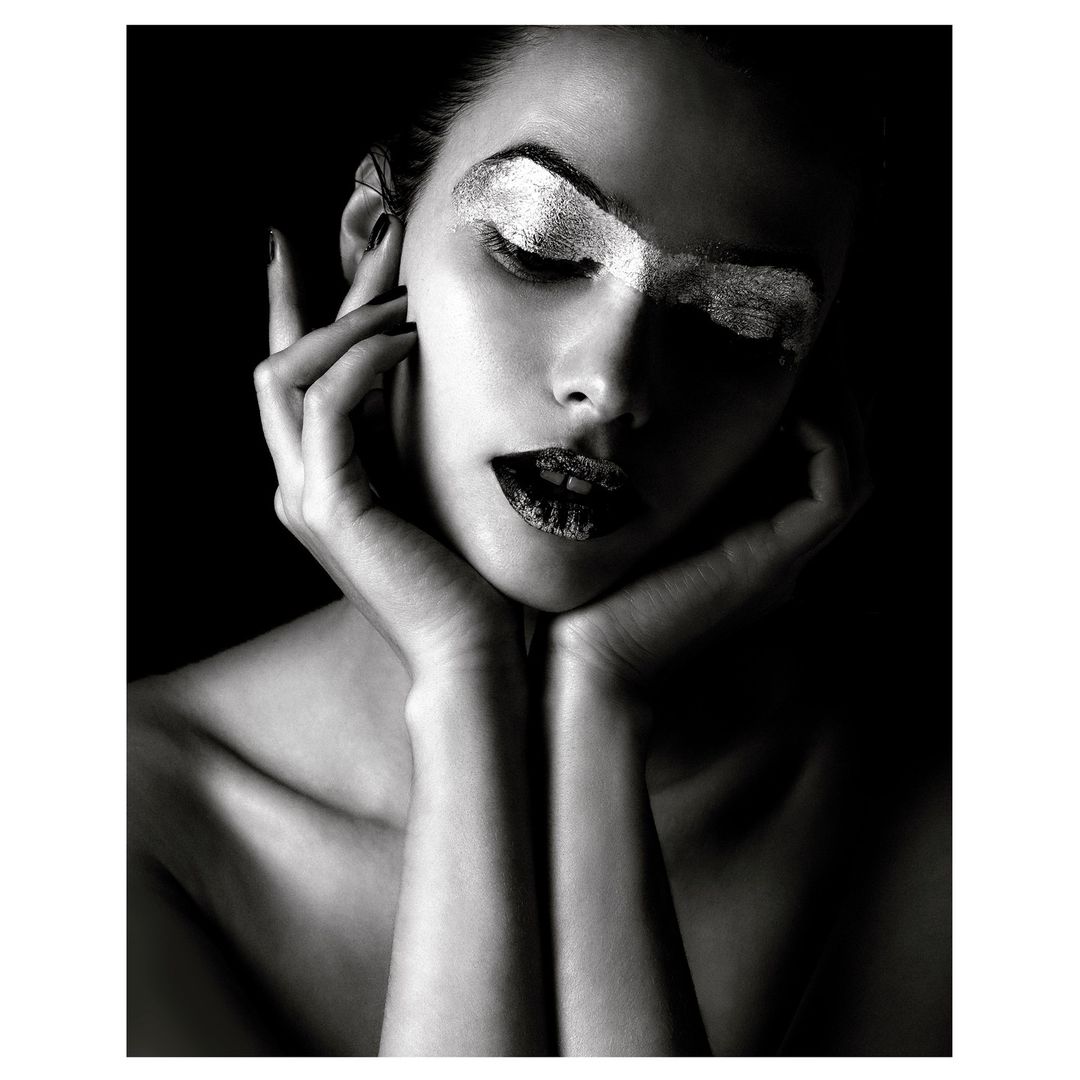 Monochrome portrait of a woman with silver eyeshadow and dark lipstick, hands framing her face, against a black background.