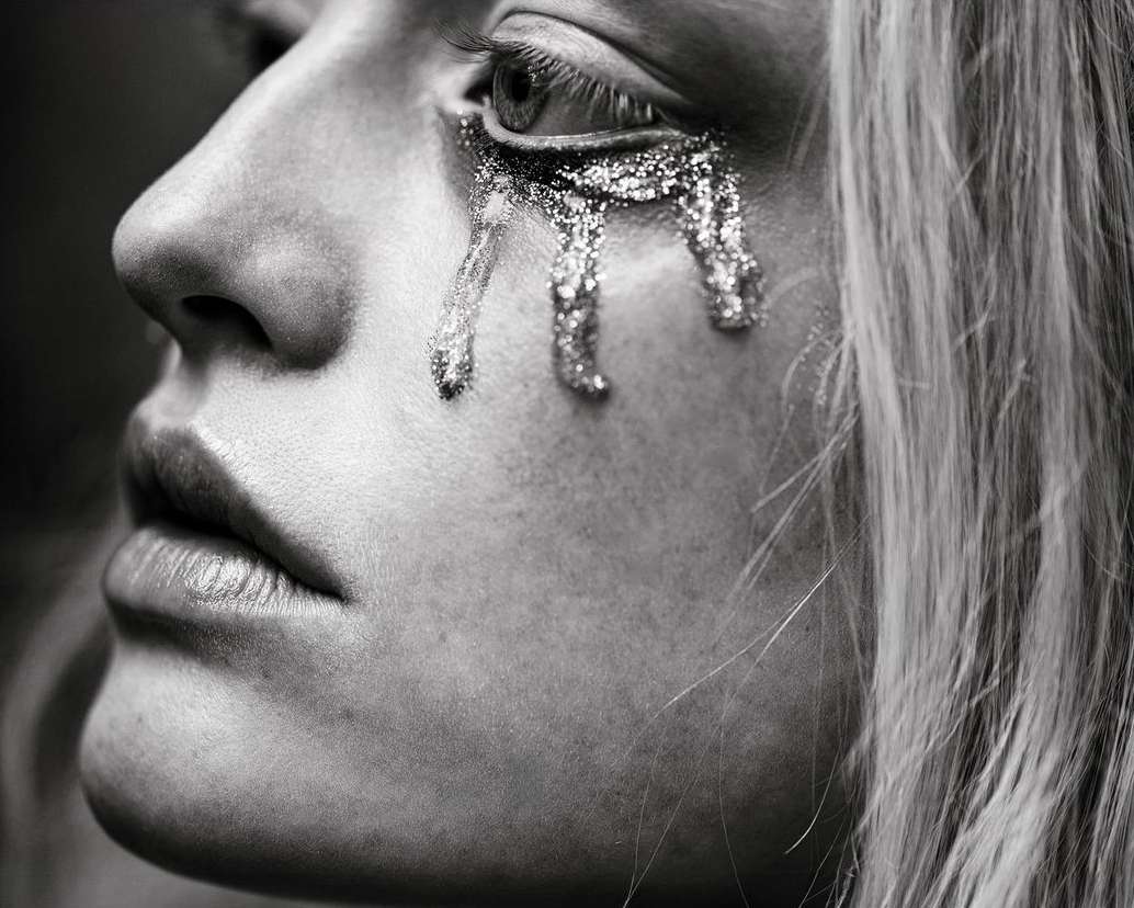 Close-up profile of a woman with metallic glitter tears dripping from her eye, photographed in black and white by Patric Shaw.