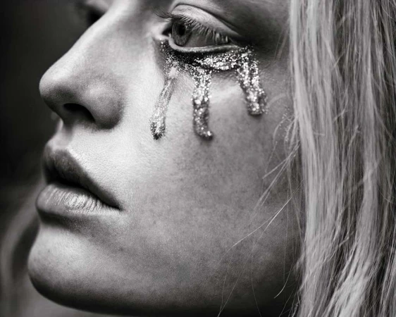 Close-up profile of a woman with metallic glitter tears dripping from her eye, photographed in black and white by Patric Shaw.