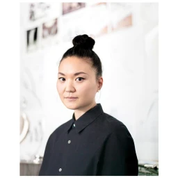 Portrait of a woman with dark hair in a top knot wearing a black buttoned shirt against a bright white background
