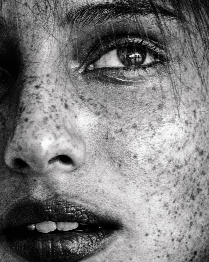 Extreme close-up portrait of a woman face in monochrome, showing detailed skin texture and prominent freckles.
