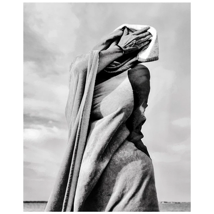 Monochrome photograph of a person wrapped in a draped towel holding their hands over their face against an overcast sky background.