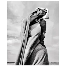 Monochrome photograph of a person wrapped in a draped towel holding their hands over their face against an overcast sky background.