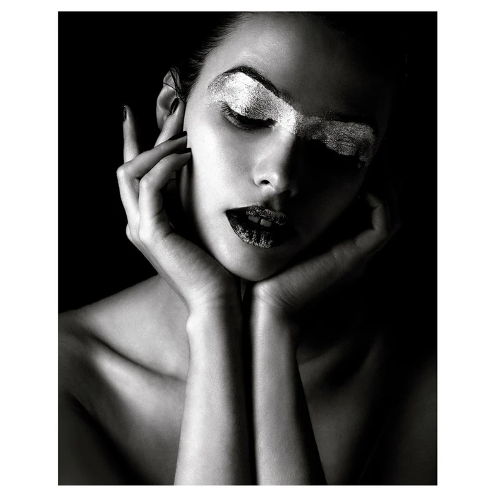Close-up portrait of a woman with metallic silver eye makeup and dark lips, framing her face with both hands against a black background.