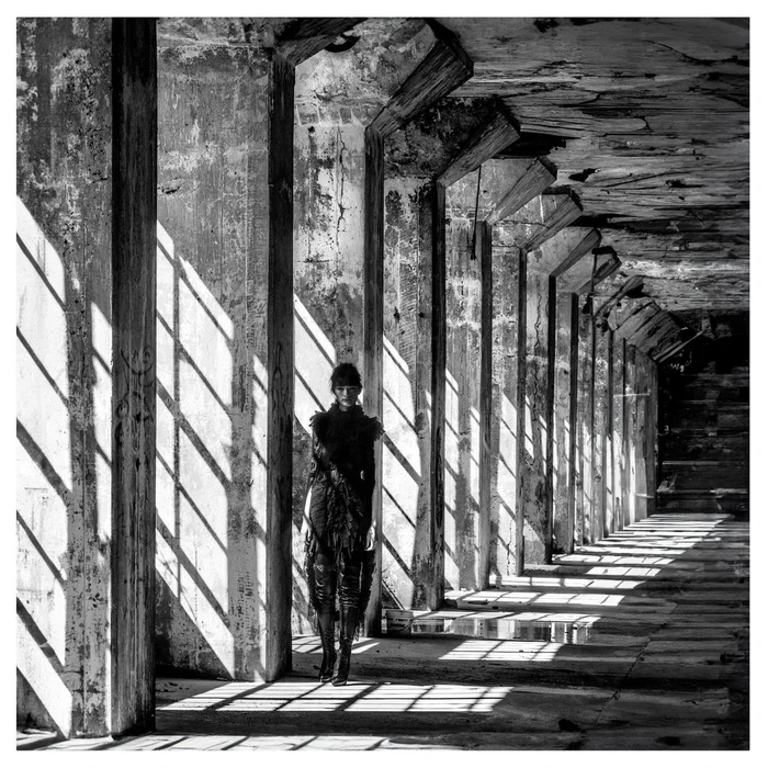 Model standing in a long weathered concrete colonnade with dramatic slanting shadows cast across the floor by Patric Shaw