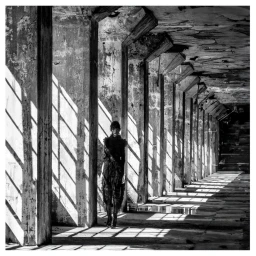 Model standing in a long weathered concrete colonnade with dramatic slanting shadows cast across the floor by Patric Shaw