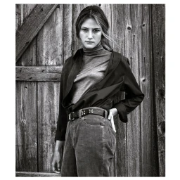 Monochrome portrait of a female model wearing a turtleneck and button-down shirt standing against a rustic wooden wall by Patric Shaw.