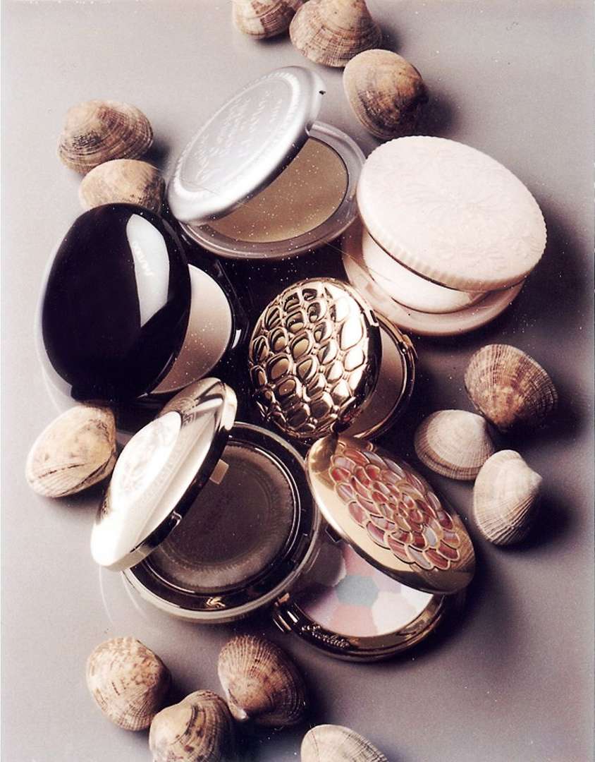 Still life arrangement of various opened and closed cosmetic compacts, with seashells surrounding them on a smooth gray surface.