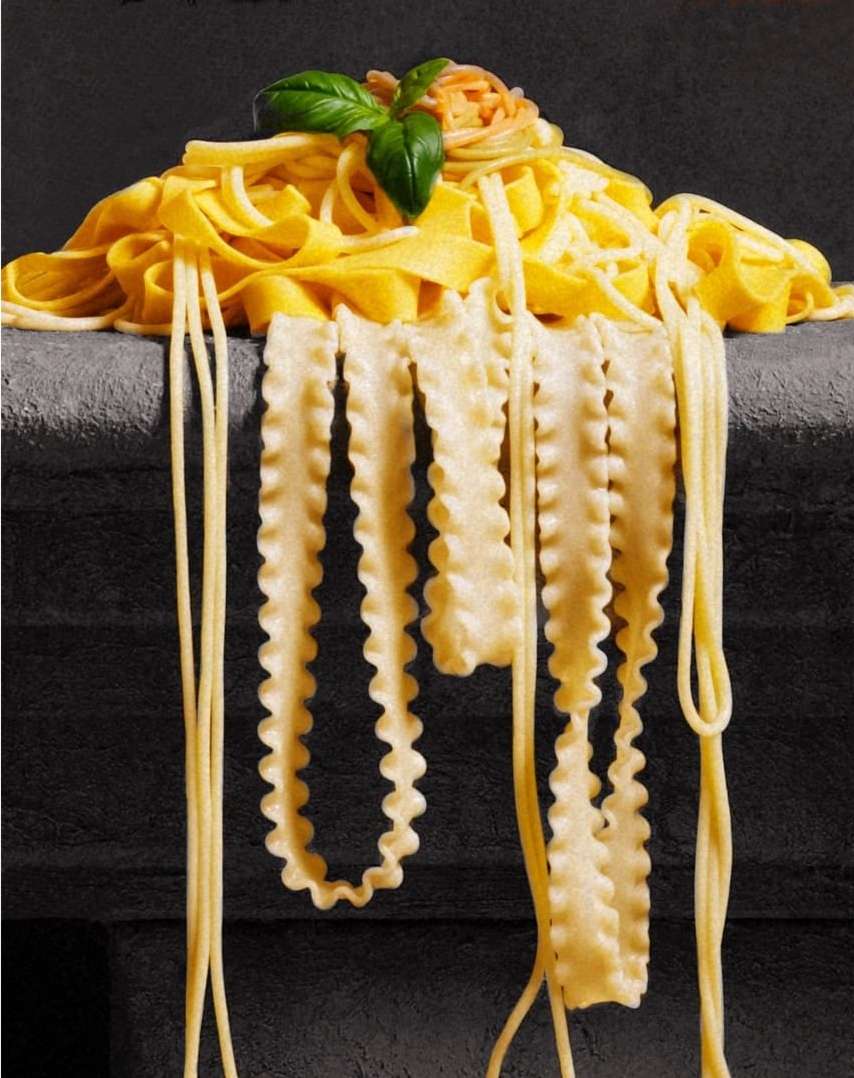 Overhead shot of a pile of fettuccine pasta and hanging strands of mafaldine pasta, topped with herbs on a stone surface.