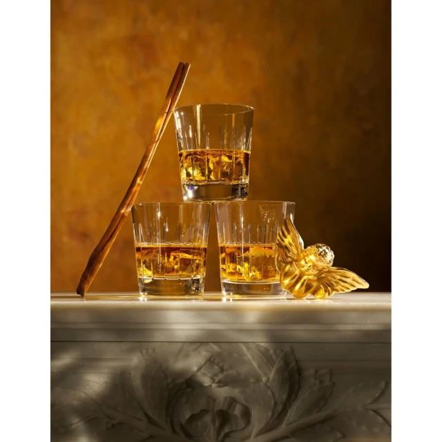 Three whiskey glasses stacked with liquid and ice, plus gold cherub and cinnamon sticks, on a marble surface against a tan background.