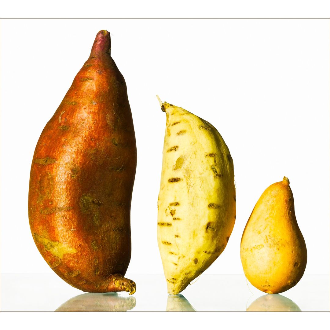 Studio shot of three sweet potatoes, varying in size and color, displayed on a reflective surface against a bright white backdrop.
