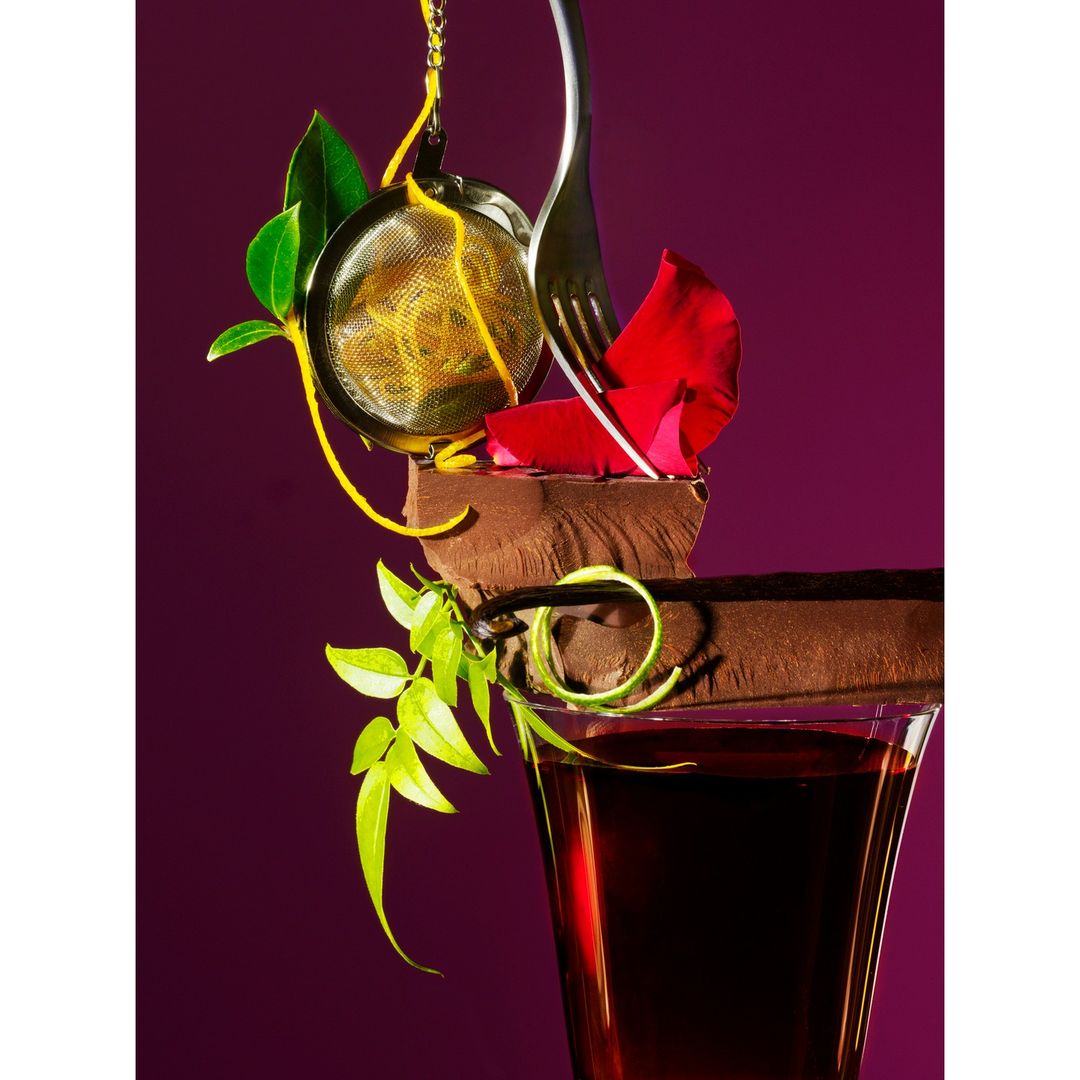 Still life of a drink with a chocolate garnish, rose petals, and lemon peel against a purple background. Fine art photography.