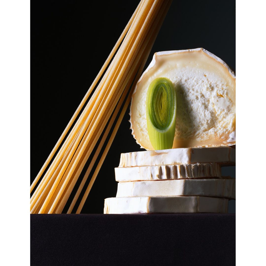 Still life photo of a stack of white cheese blocks, a green leek slice, and a bundle of spaghetti against a black background.