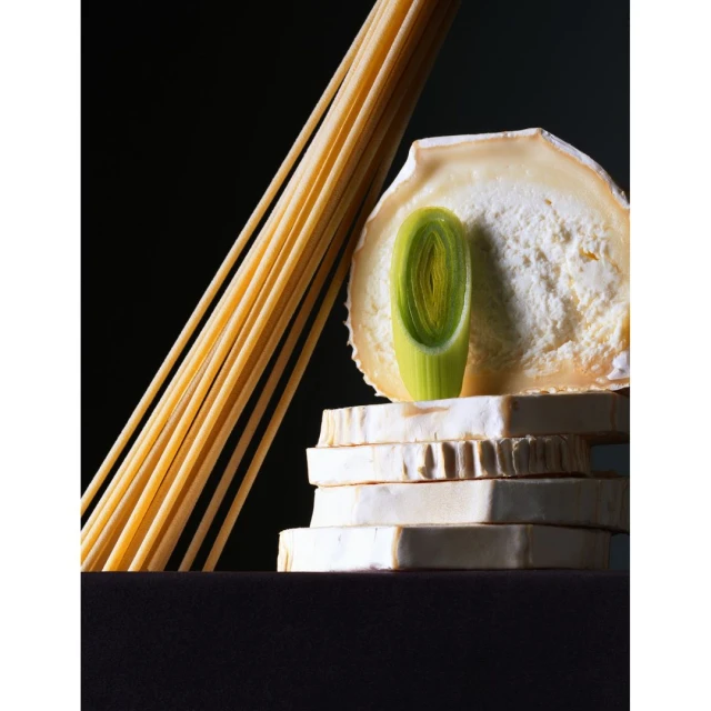 Still life photo of a stack of white cheese blocks, a green leek slice, and a bundle of spaghetti against a black background.