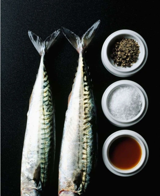 Overhead shot of two raw mackerels with seasonings, salt, pepper, and soy sauce, set against a dark backdrop for food photography.