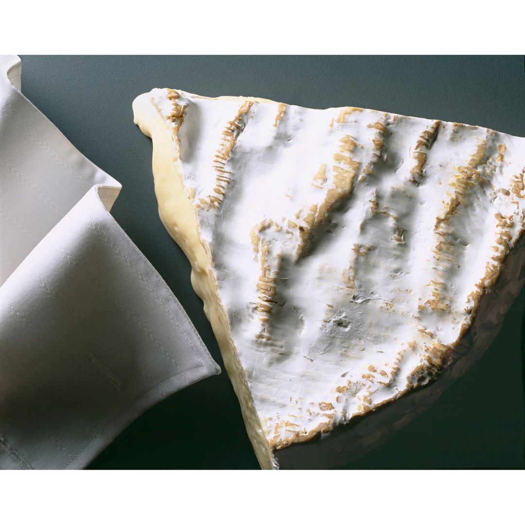 Close-up of a wedge of white mold cheese next to a white folded cloth, set against a dark green background, indoor studio food photography.