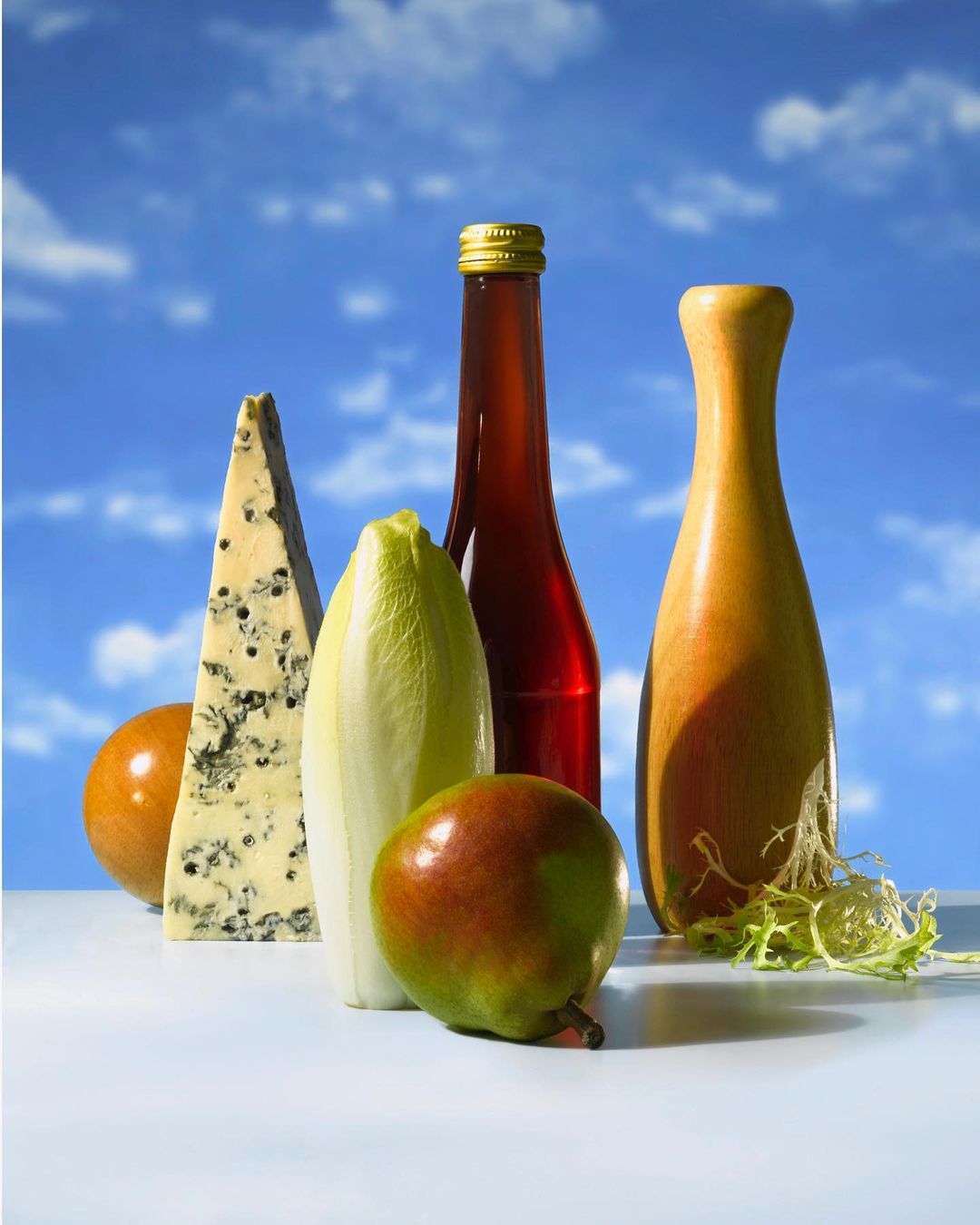 Still life composition of gourmet food items including blue cheese, a pear, endive, salad greens, and two bottles against a blue sky.