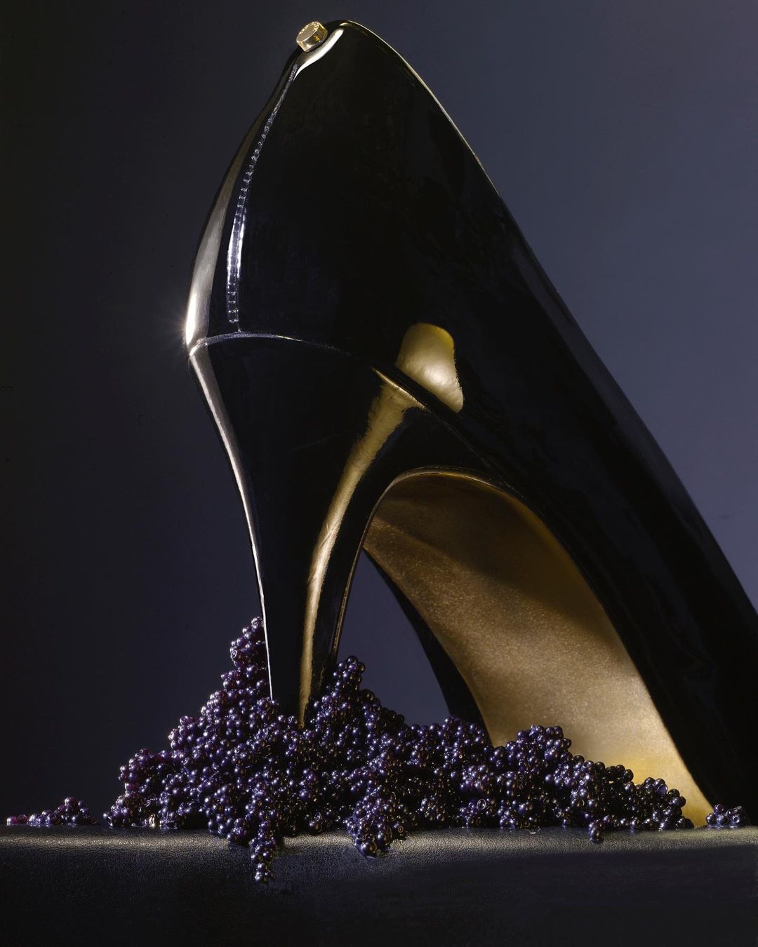 Elegant black patent leather high heel shoe presented on dark backdrop, adorned with scattering of lustrous black caviar beads.