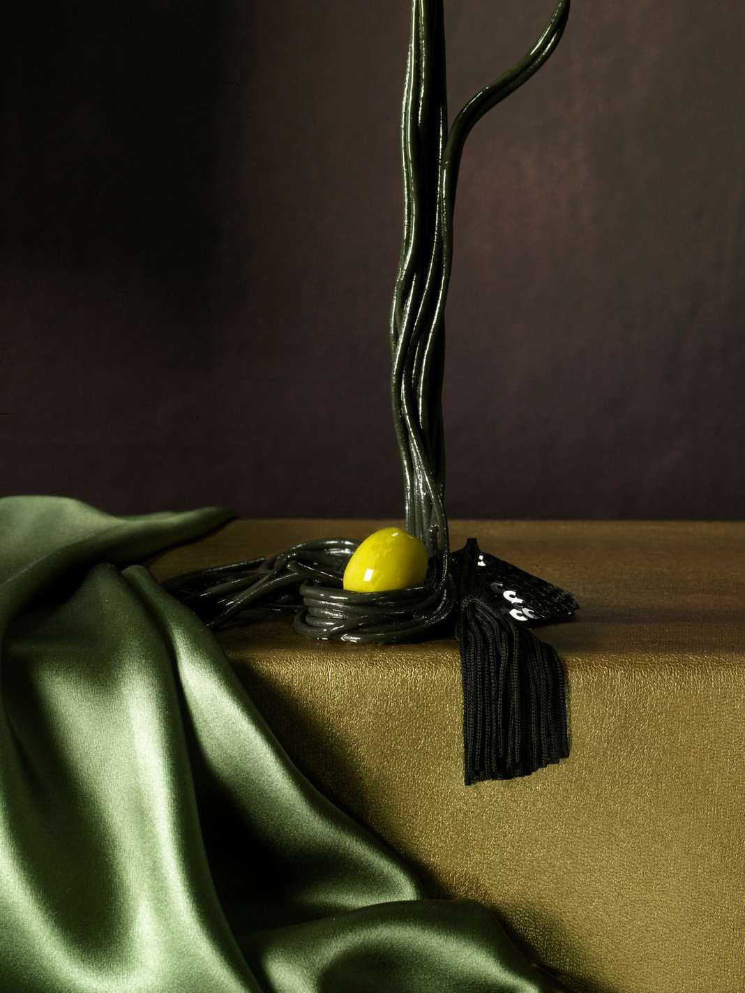 Abstract still life photography of a dark metallic tree sculpture with a yellow egg-shaped object and draped green satin fabric.