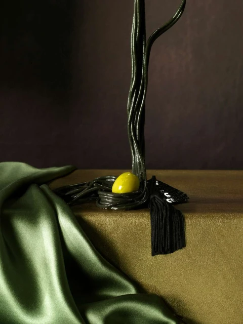 Abstract still life photography of a dark metallic tree sculpture with a yellow egg-shaped object and draped green satin fabric.