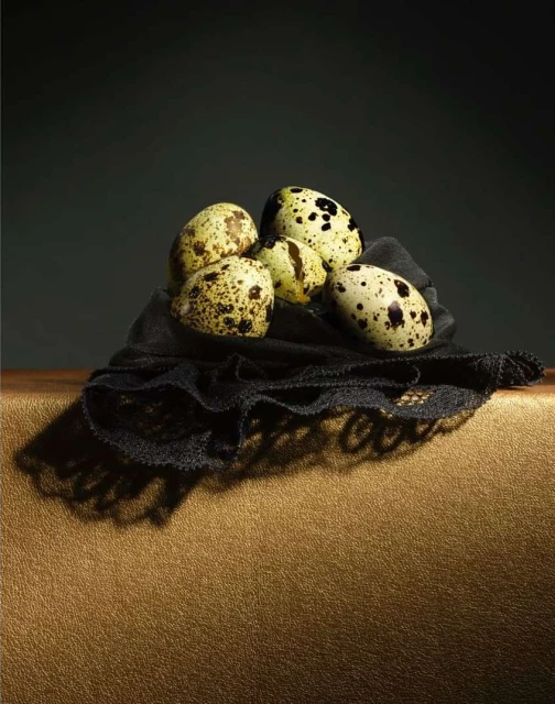 Still life photograph of five speckled quail eggs arranged on a draped black lace cloth, set against a moody, dark backdrop.