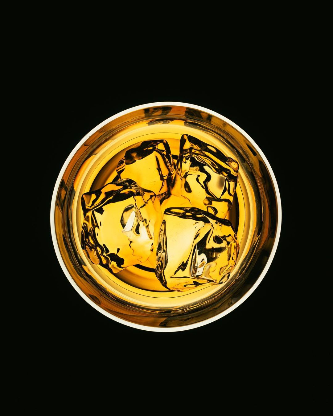 Overhead shot of a clear glass filled with golden liquor and ice cubes, set against a stark black background, enhancing the drink's rich color.