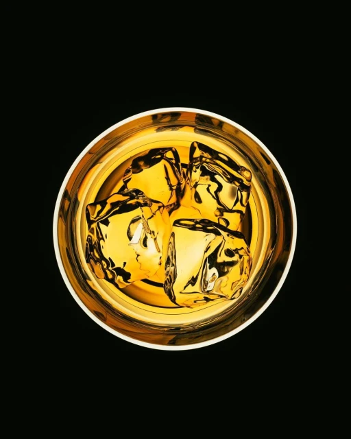 Overhead shot of a clear glass filled with golden liquor and ice cubes, set against a stark black background, enhancing the drink's rich color.