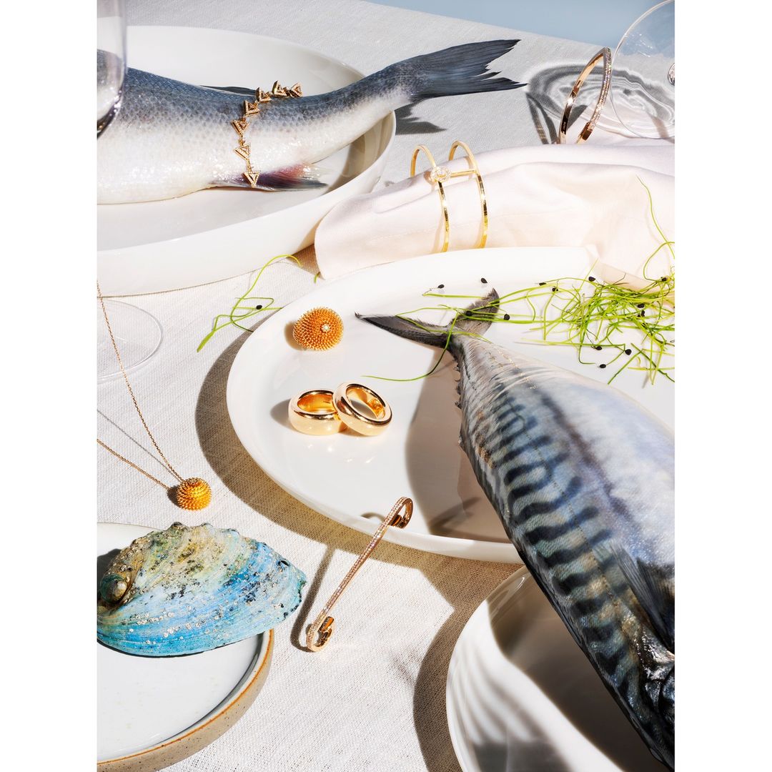 Overhead still life photograph of plates with fish, jewelry, and decorative elements set against a white tablecloth backdrop.