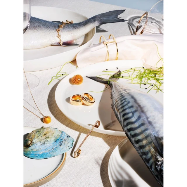 Overhead still life photograph of plates with fish, jewelry, and decorative elements set against a white tablecloth backdrop.