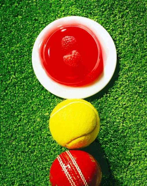 Overhead shot of red gelatin with raspberries on a plate, a tennis ball, and cricket ball on green grass.