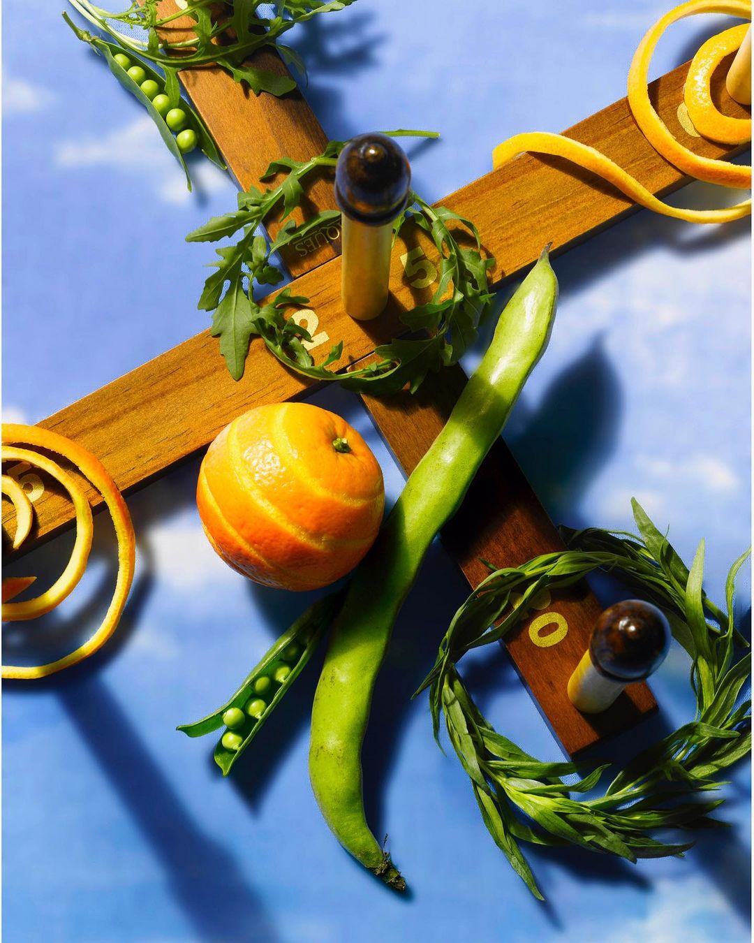 Overhead food photography of an orange, green beans, peas in a pod, and citrus peel arranged on a wood cross against a blue background.