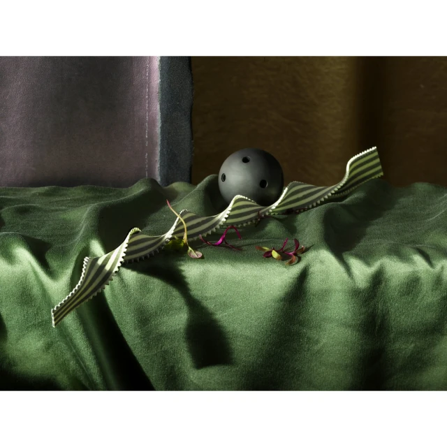 Stripped ribbon and dark ball on wrinkled green satin with green shoots, set against a purple panel and golden wall in moody lighting.