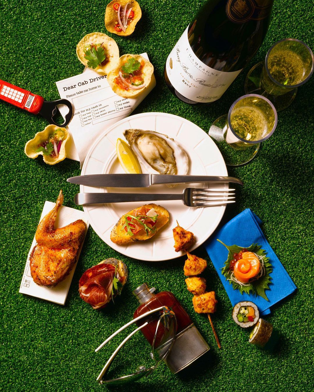 Overhead picnic flatlay with champagne bottle, oysters, sushi, snacks, cutlery, and drinkware arranged on green turf background.