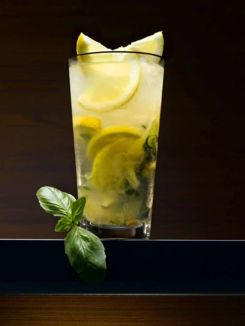 Tall glass filled with a cool, refreshing mojito drink, garnished with lemon slices and fresh basil leaves against a dark backdrop.