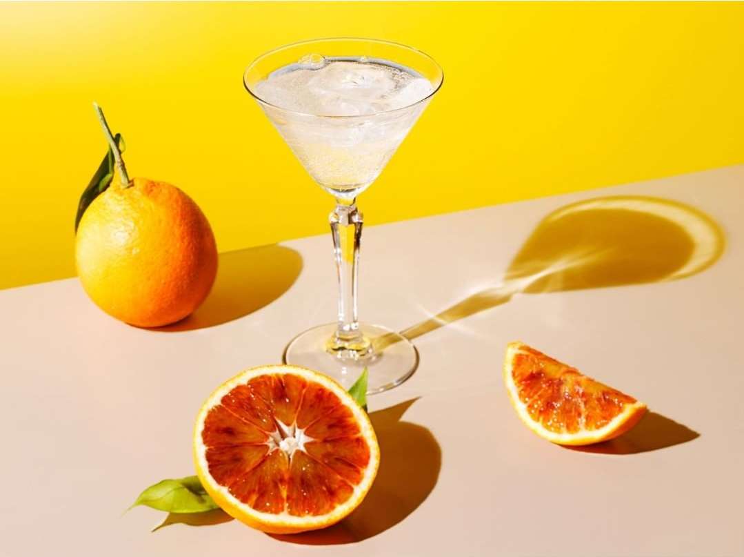 Brightly lit image of a martini glass filled with liquid and ice, surrounded by whole and sliced blood oranges against a yellow backdrop.
