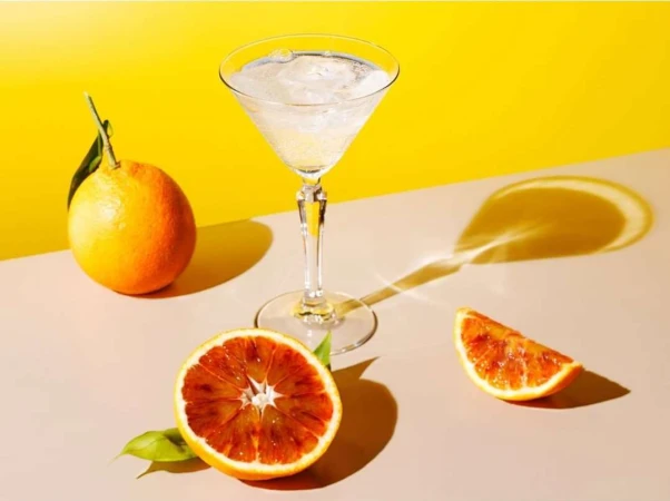Brightly lit image of a martini glass filled with liquid and ice, surrounded by whole and sliced blood oranges against a yellow backdrop.