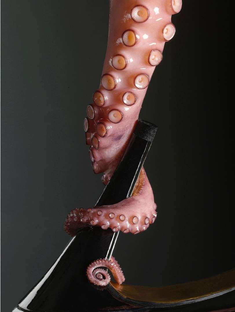 Octopus tentacle clings to a glossy black high heel shoe against a dark background in a studio product shot with dramatic lighting.