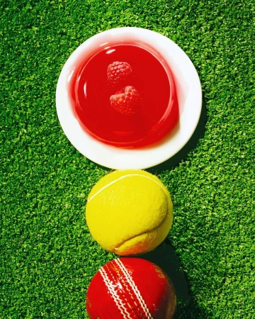 Overhead shot of red jello with raspberries, tennis ball, and cricket ball on artificial green turf background, bright and colorful.