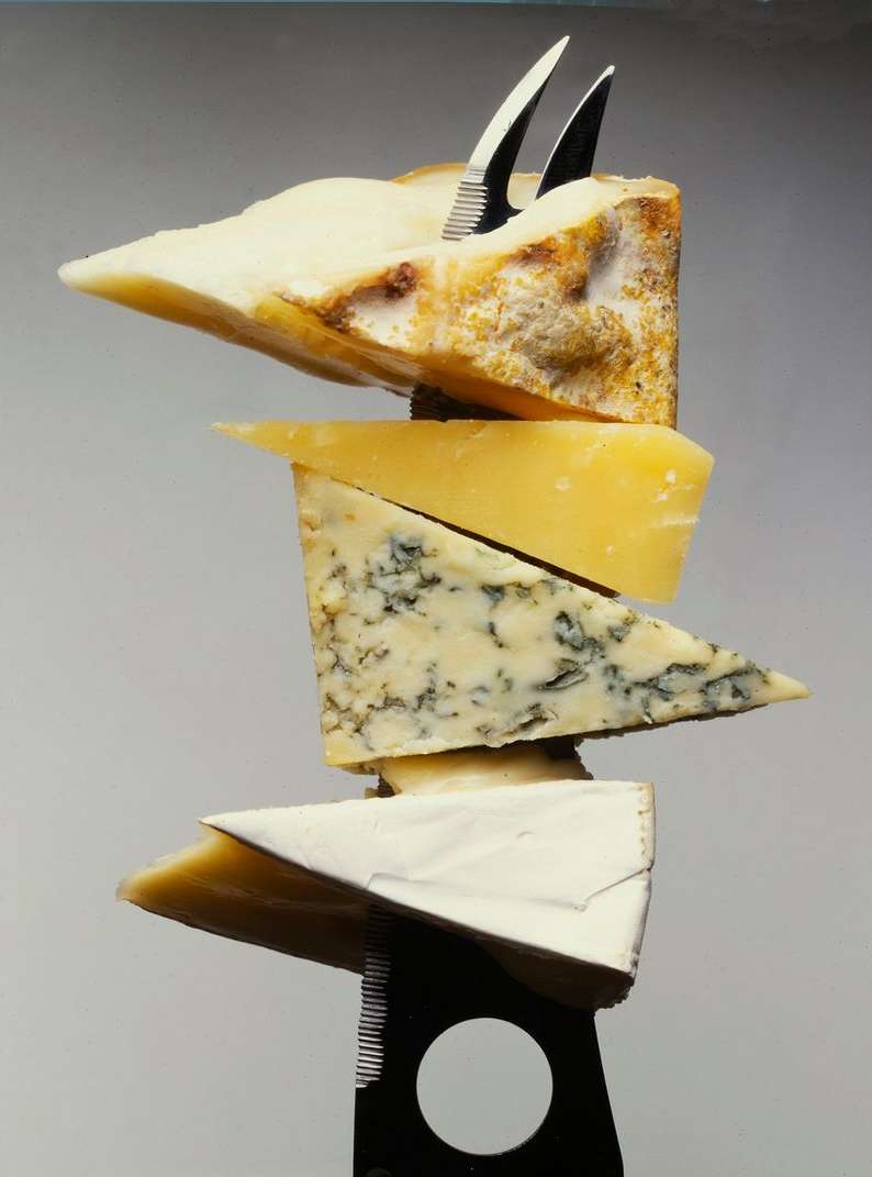Tower of various cheeses stacked on top of each other, pierced by a cheese knife. Food photography of cheese and cutlery.