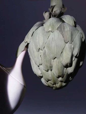 Close-up still life of a fresh artichoke leaning against a pale pitcher with a dark purple background, studio lighting.