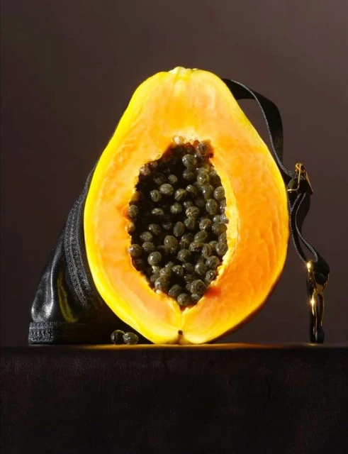 Cross section of a papaya with dark seeds in front of a handbag against a dark backdrop, showcasing a blend of nature and fashion.