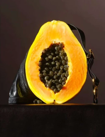 Cross section of a papaya with dark seeds in front of a handbag against a dark backdrop, showcasing a blend of nature and fashion.